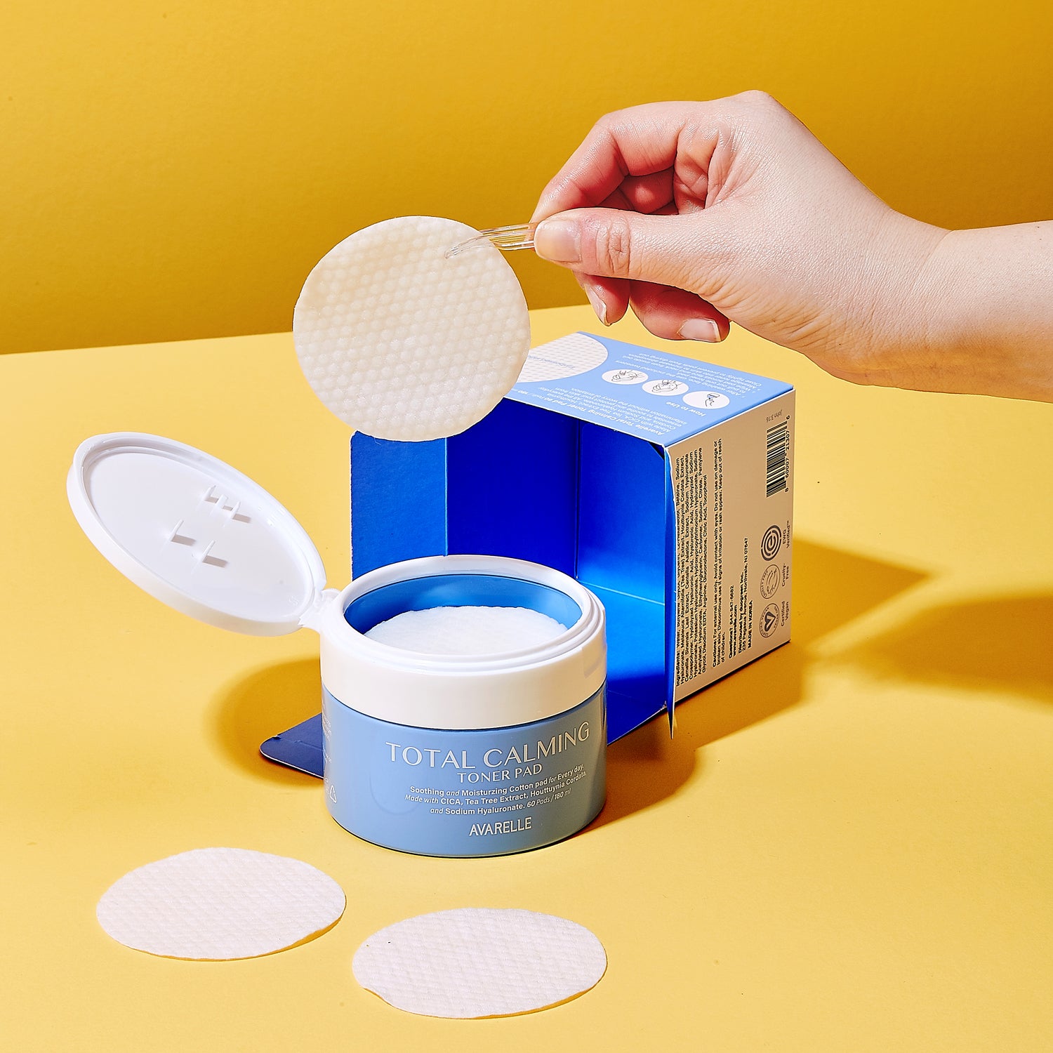 A hand holds a one of Avarelle's Total Calming Toner pads with a tweezer above an open jar of Avarelle's Total Calming Toner Pads. That contains AHA & PHA exfoliating toner pads next to its box on a yellow surface.