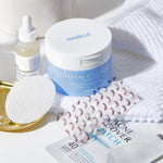 Assorted skincare products including Avarelle Total Calming Toner Pads, and ZitOut Original 40 acne cover patches, and a serum, arranged neatly on a bright surface with white towels.